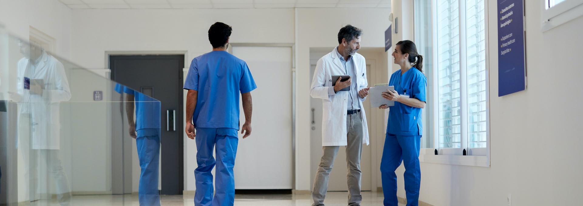 Symbolbild für Zertifizierung: Arzt und Pflegerin in Fallbesprechung auf einem Krankenhausgang, daneben ein männlicher Pfleger mit dem Rücken zur Kamera